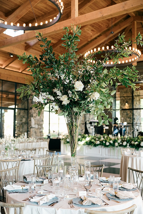 Reception tablescape with tall wedding centerpiece of white flowers and greenery in a clear vase, candles on a round table under ring chandeliers