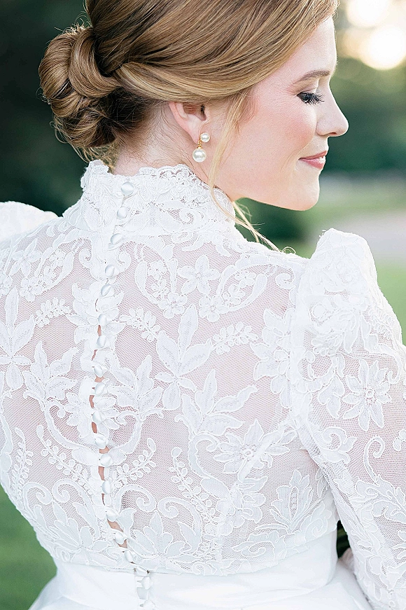 Bridal hairstyle in a low bun wedding hair updo, side profile with pearl drop earrings and lace high neck dress in blurred garden greenery