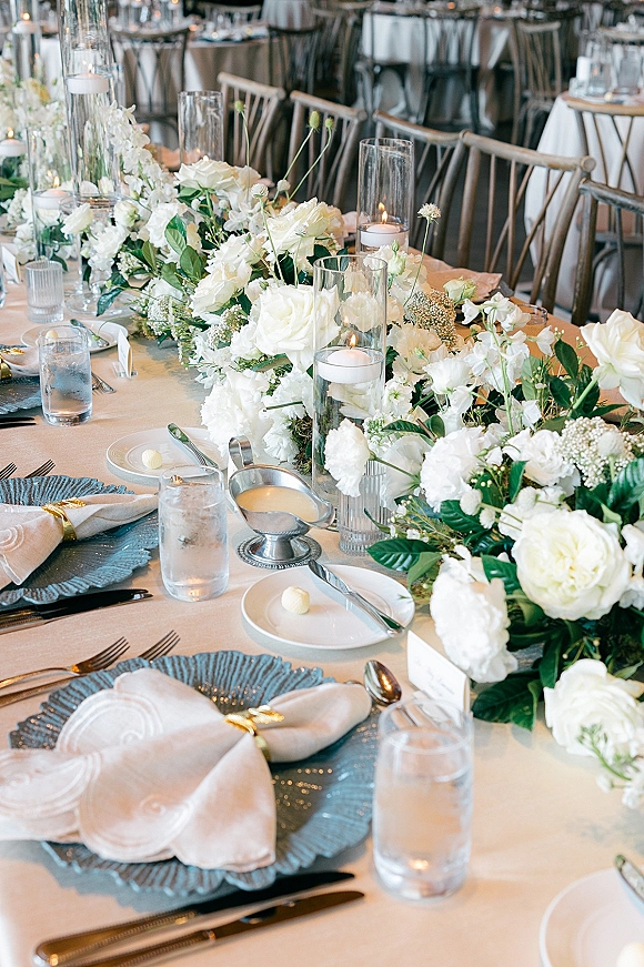 Reception tablescape with a white floral table runner and greenery garland, cylinder vases holding floating candles, elegant place settings on round tables