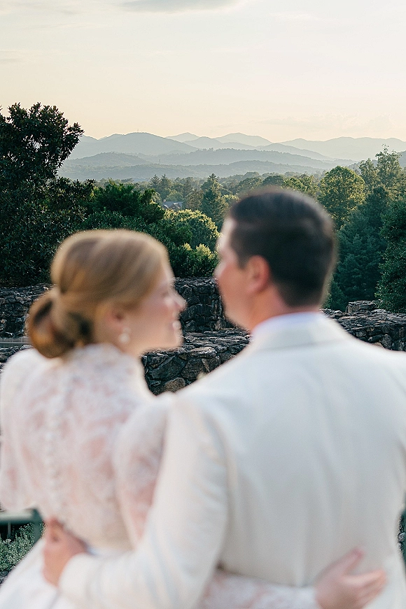 Couple portrait at a mountain wedding overlook, bride in a lace long sleeve dress and groom in a beige suit facing the view