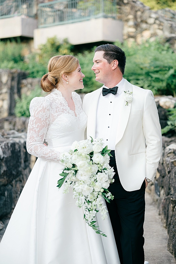 Couple portrait of bride and groom smiling, bride holding a cascading white bouquet in front of a stone wall and greenery outdoors