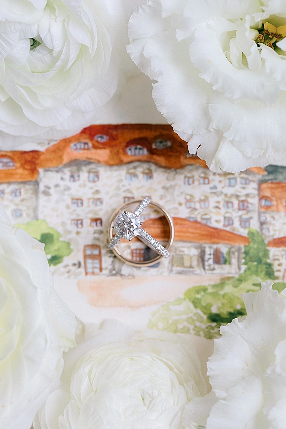 Wedding rings with a diamond engagement ring stacked on an illustrated card beside white flowers on a white surface flat lay