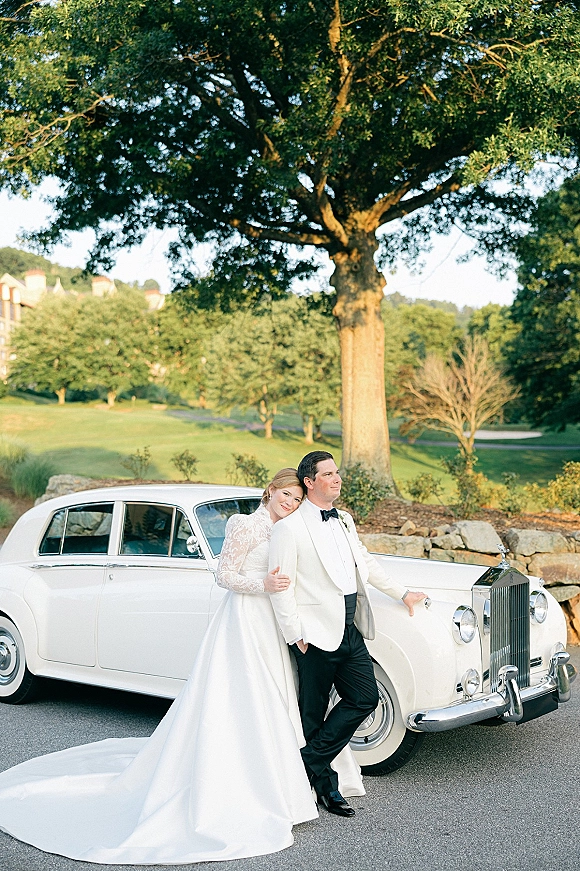 Couple portrait of bride and groom with vintage car, her long-sleeve gown and train beside his white dinner jacket on a green lawn