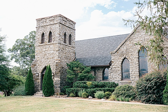 Stone church exterior with arched stained glass windows and steeple tower, front steps and lanterns, set on a manicured lawn under clouds