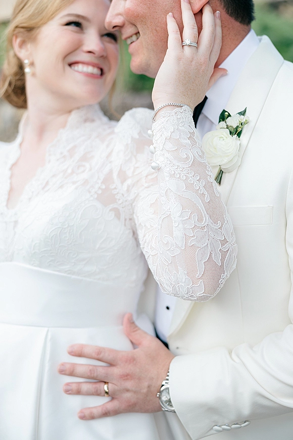 Wedding couple portrait of bride and groom close up, her hand on his face showing lace sleeve and rings against blurred greenery backdrop