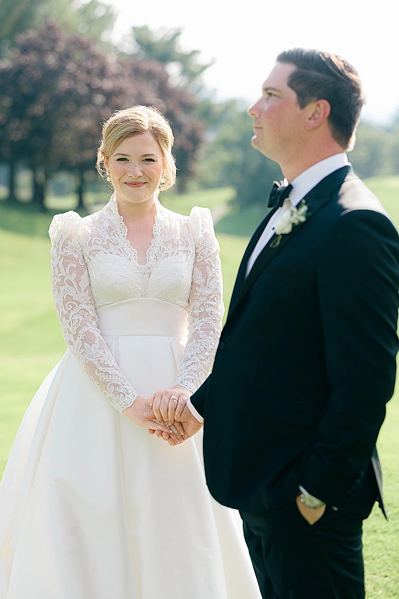 Couple portrait of bride and groom holding hands, bride in long sleeve lace dress beside tuxedoed groom on a green lawn in daylight