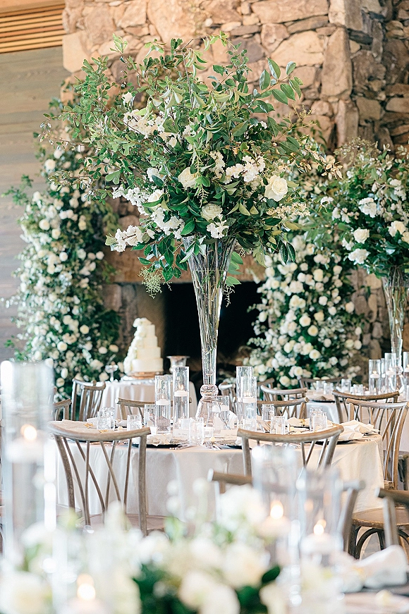 Reception tablescape with tall greenery centerpiece, white flowers and candles on round white linen tables, crossback chairs by stone fireplace