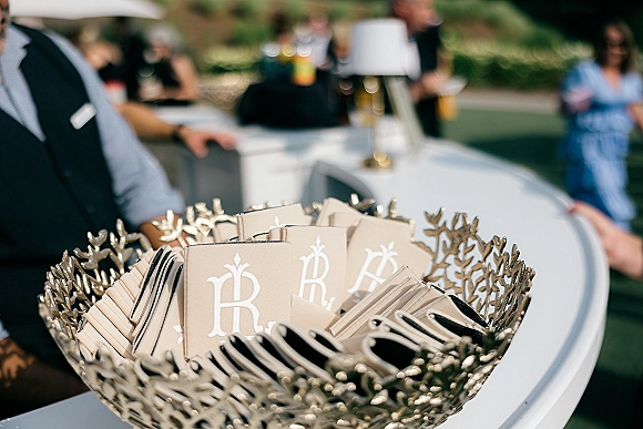Wedding favors in monogram wedding favors packets with tags arranged in a metal leaf bowl on a white bar counter during outdoor cocktail hour