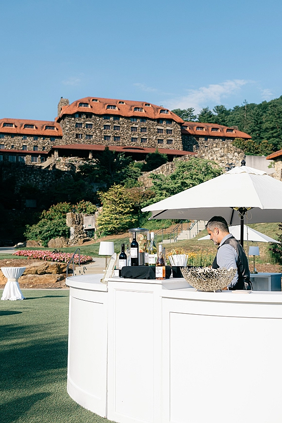 Outdoor wedding bar wedding bar setup with a white counter, liquor and wine bottles, drink dispenser, and bar umbrella on a lawn by a stone lodge