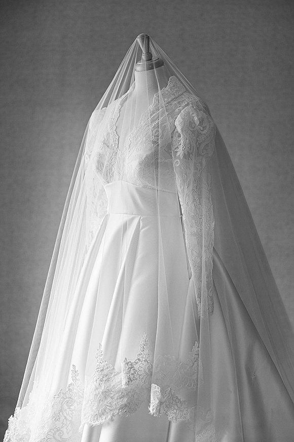 Wedding dress with lace sleeve details on a mannequin, featuring an illusion lace bodice, satin skirt, and cathedral veil against a plain wall