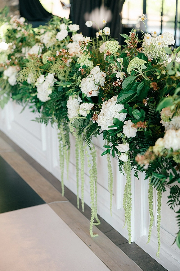 Wedding floral installation with white flowers and green foliage, hydrangeas and daisies on a runner against a white paneled wall in window light