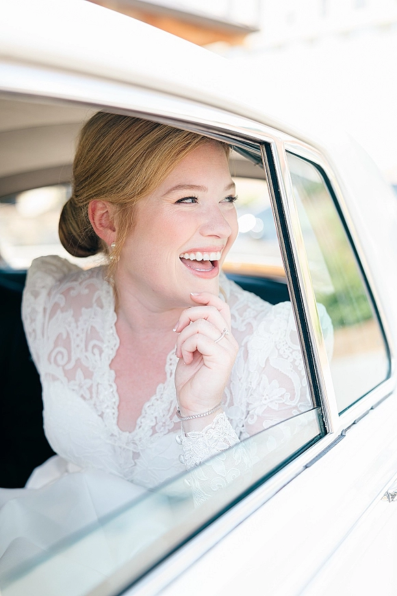 Bridal portrait of a bride in car laughing by the window in a lace wedding dress, pearl earrings visible in daylight inside a vintage car