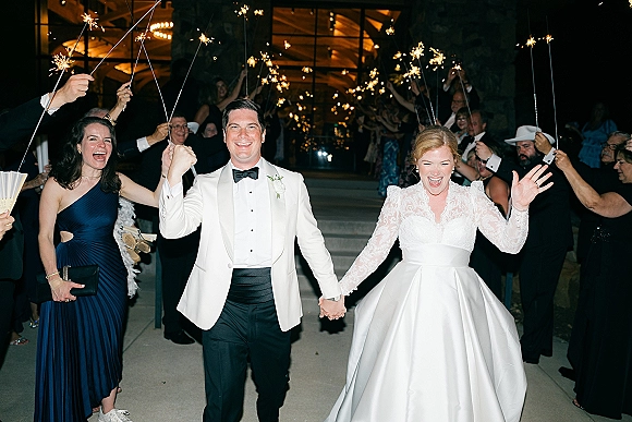 Wedding send-off with a sparkler exit wedding as bride in lace sleeves waves beside groom in tuxedo at venue entrance at night with guests