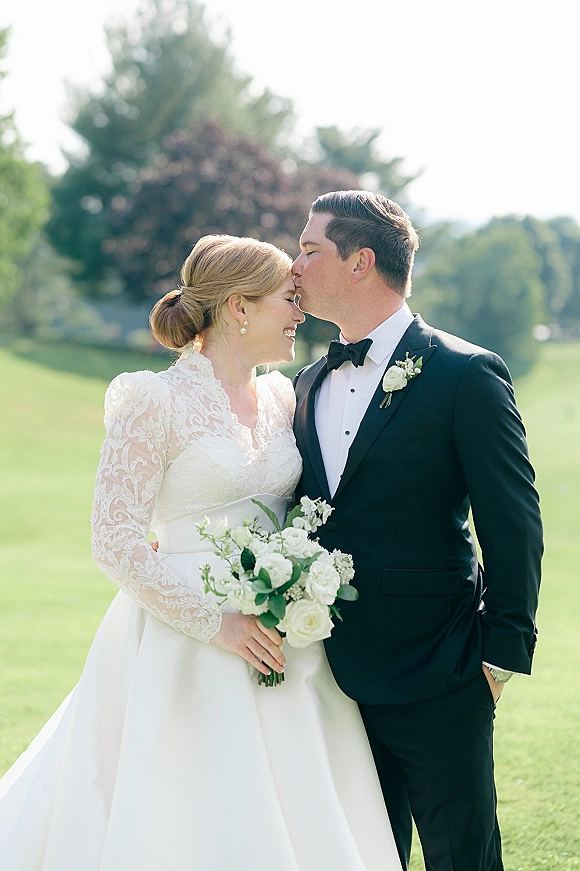 Wedding couple portrait of groom kissing bride’s forehead as she holds a white rose bouquet, standing on a green lawn with trees behind