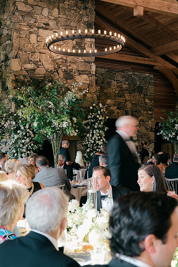 Reception decor with greenery garland and white florals framing a wedding cake on a stone fireplace, candlelit beneath a round chandelier