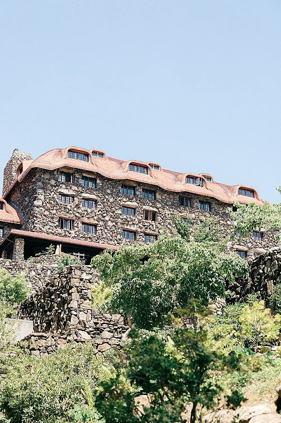 Stone lodge exterior with copper roof and dormer windows, framed by trees and shrubs under a clear blue sky on a hillside