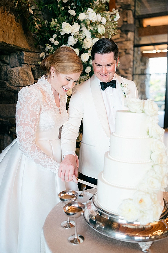 Wedding cake cutting as bride in lace long sleeves and groom in white tux cut a tiered cake with white roses by a stone wall