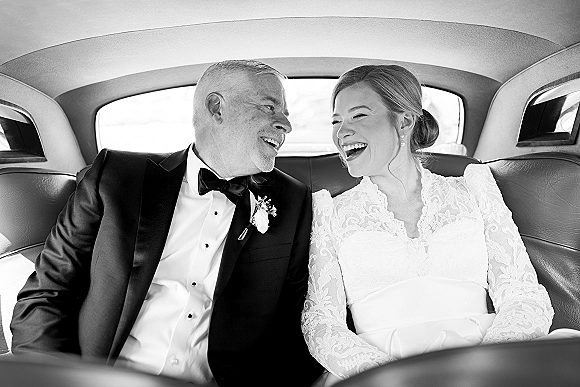 Couple portrait in a black and white wedding photo, bride in long sleeve lace dress and groom in tuxedo laughing in a leather backseat