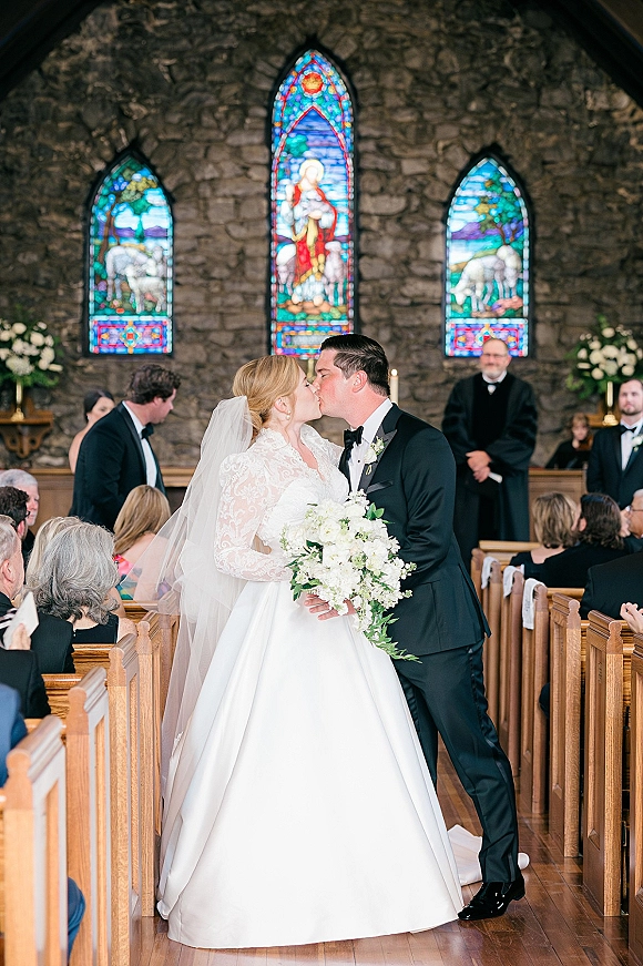 Wedding kiss as bride and groom share their first kiss wedding ceremony in a church aisle, bride holding a bouquet before stained glass windows
