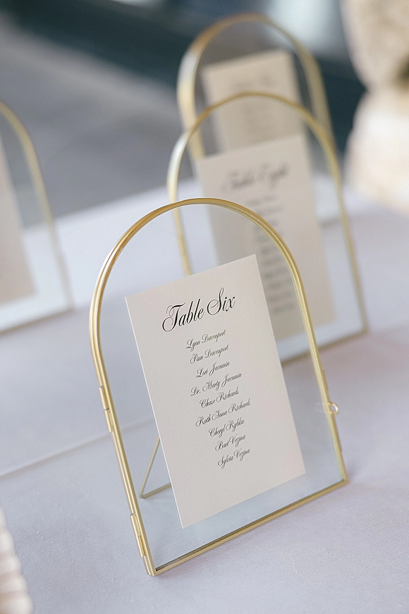 Wedding table numbers in gold table number frames with a calligraphy card on a white tablecloth, blurred reception tables behind