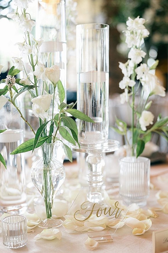 Reception tablescape with wedding table centerpiece of white flowers in glass vases, tall cylinder and floating candles, bokeh lights behind