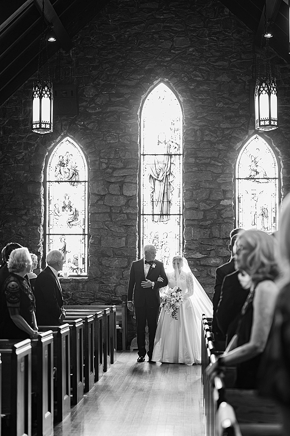 Wedding processional with bride walking down aisle in a ballgown and cathedral veil, holding a bouquet beside a tuxedoed escort in a church aisle with pews and stained glass windows