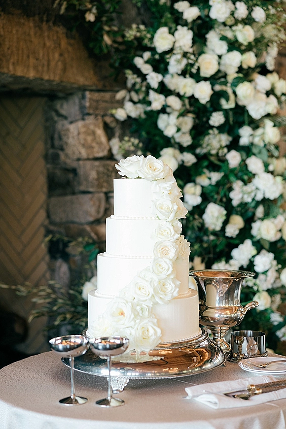 Wedding cake with roses, a white tiered design on a silver tray, set against a greenery and white rose floral wall backdrop