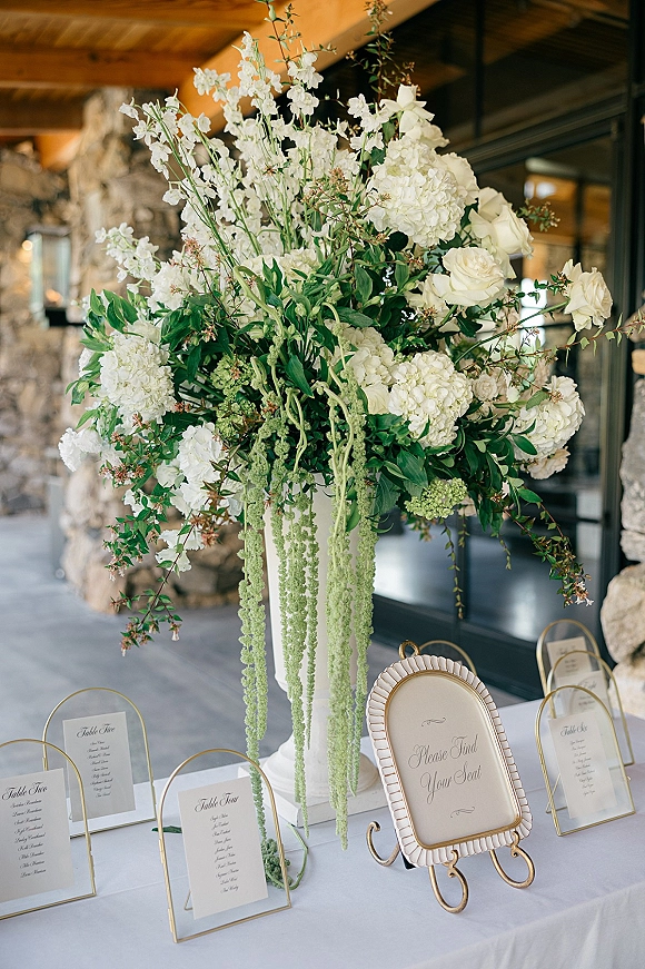 Escort card table with wedding seating chart display in gold arch frames, topped with tall white rose and hydrangea centerpiece on a pedestal vase
