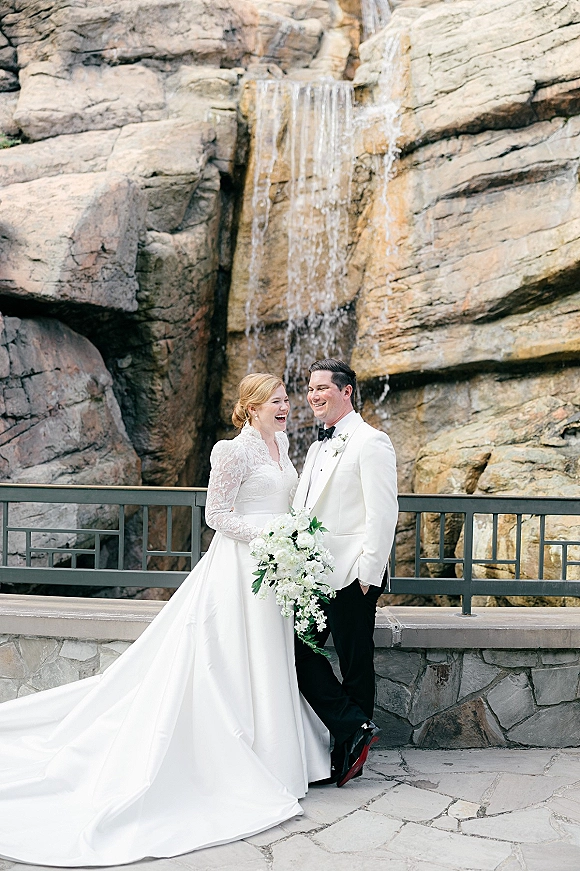 Couple portrait in an outdoor wedding portrait, bride in long sleeve lace gown holding a cascading bouquet beside groom in tux at a waterfall terrace