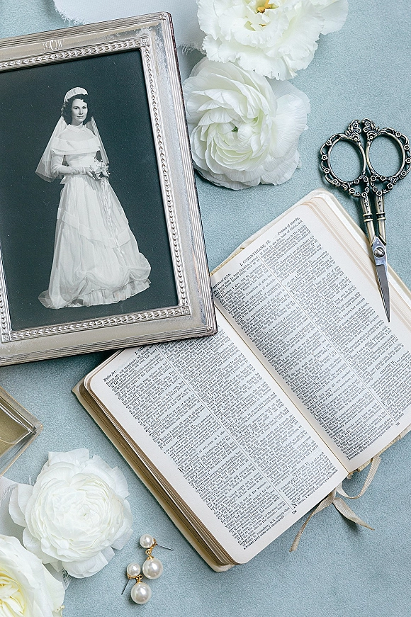 Wedding flat lay with a bridal heirloom flat lay of framed bridal photo, open book, white flowers, pearl earrings and ornate scissors on blue fabric