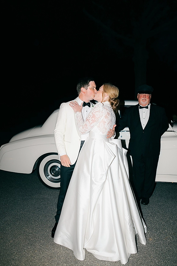 Wedding kiss as bride in lace-sleeved satin ballgown and groom in white jacket embrace by a vintage white car at night