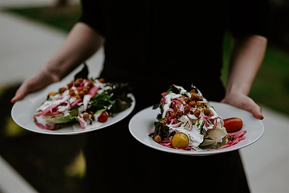 Wedding catering plates of mixed greens salad with pickled onions, cherry tomatoes, and creamy dressing on white plates outdoors by greenery