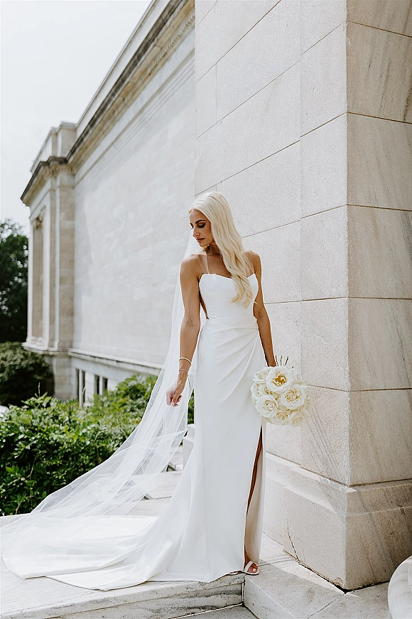 Bridal portrait of a bride in a strapless wedding dress with long veil, holding a white rose bouquet on stone steps by columns