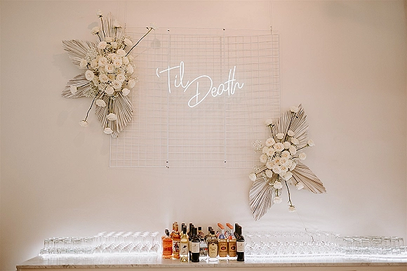 Bar backdrop decor with a wedding bar setup, wire grid and neon sign accented by white roses, dried palm leaves, and liquor bottles on a lit counter