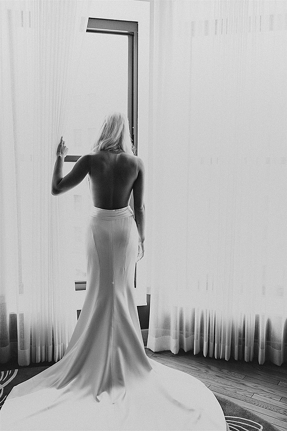 Bridal portrait of a bride by window in a strapless low back gown with a long train, standing on a hotel room wood floor by sheer curtains