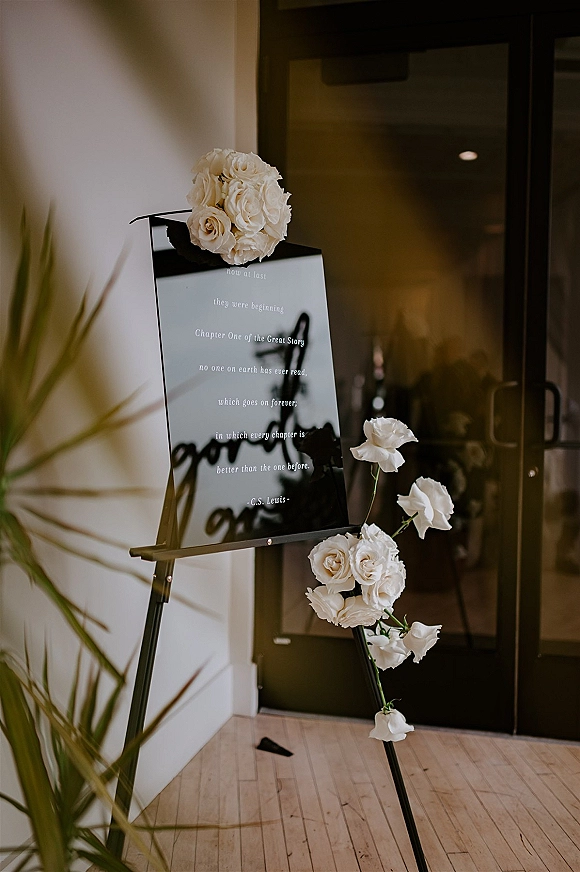 Wedding welcome sign on an easel, acrylic panel with white rose arrangement at an indoor entryway with glass doors and wood floor