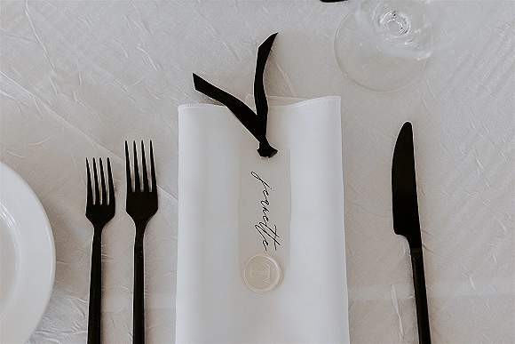 Wedding place setting with a minimalist wedding place setting featuring a white napkin, calligraphy place card, black ribbon and wax seal on white tablecloth