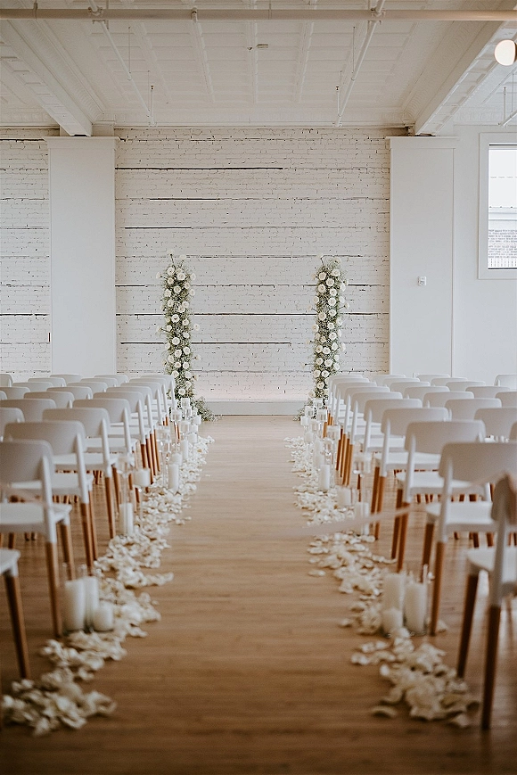 Ceremony aisle design with white floral pillars and petal-lined runner, candle aisle markers and chairs in a bright white-brick room