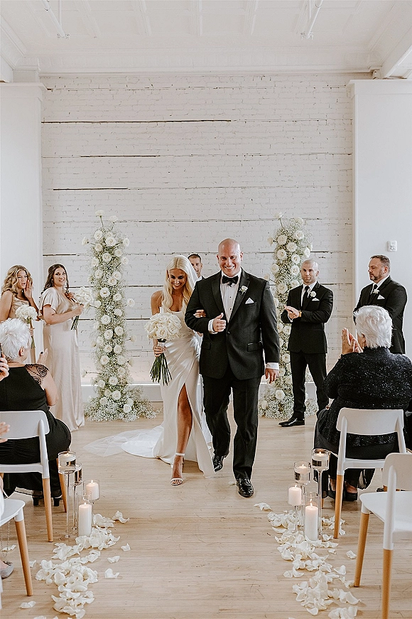 Wedding recessional as bride and groom walk the aisle past candlelit glass holders and rose petals, framed by white brick wall and floral pillars
