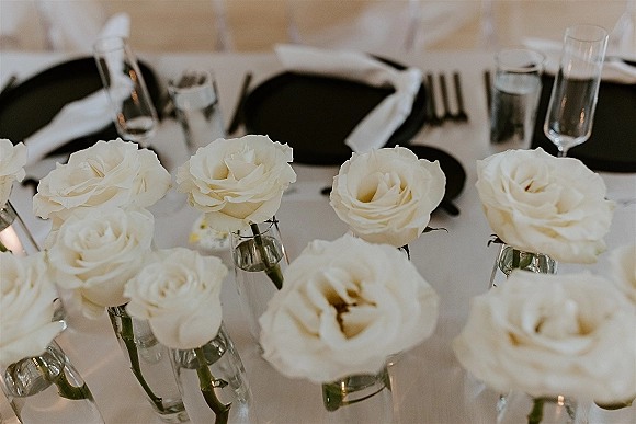 Wedding tablescape with white rose centerpieces in bud vases, black dinner plates, silver flatware, champagne flutes, and votive candles