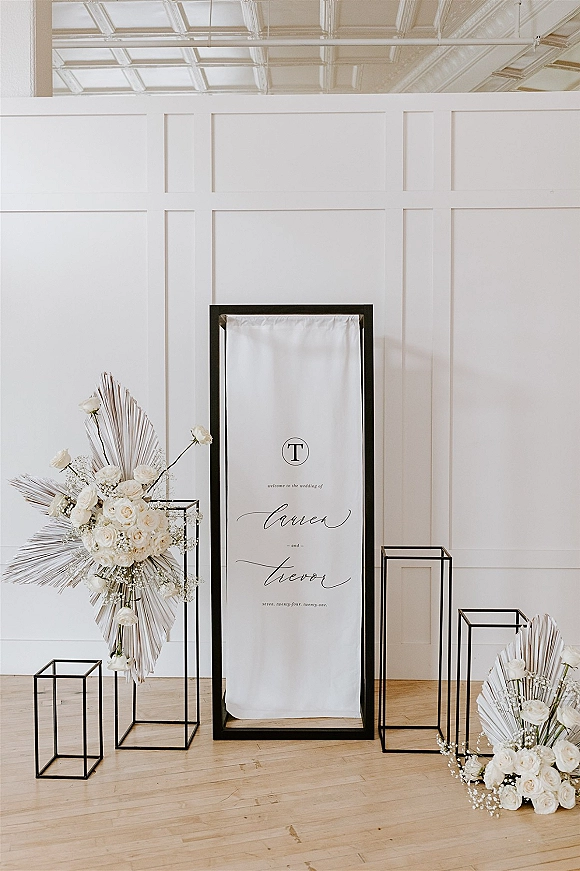 Wedding welcome sign in a black frame with white roses and baby’s breath on acrylic box pedestals against a white paneled wall indoors