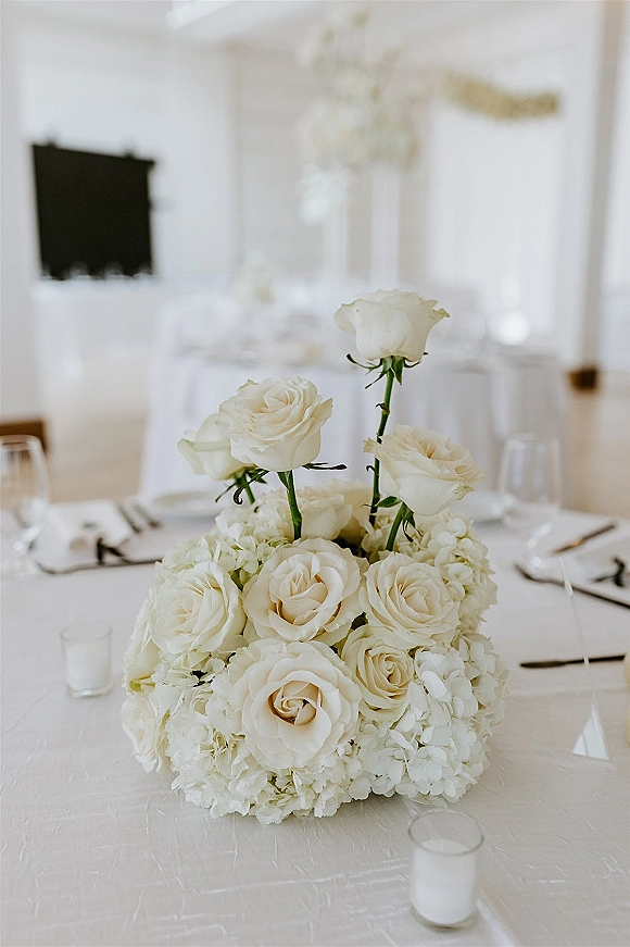 Wedding centerpiece with white rose centerpiece and hydrangeas, surrounded by wine glasses and votive candles on a white tablecloth indoors