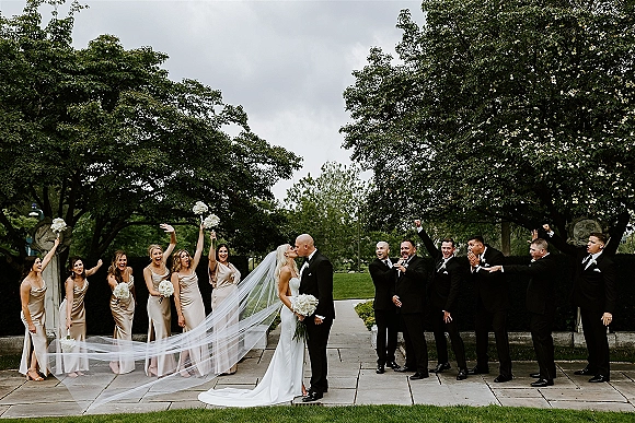 Wedding party photo of bride and groom kiss as bridesmaids and groomsmen cheer, bride holding white bouquet on a stone walkway in gardens