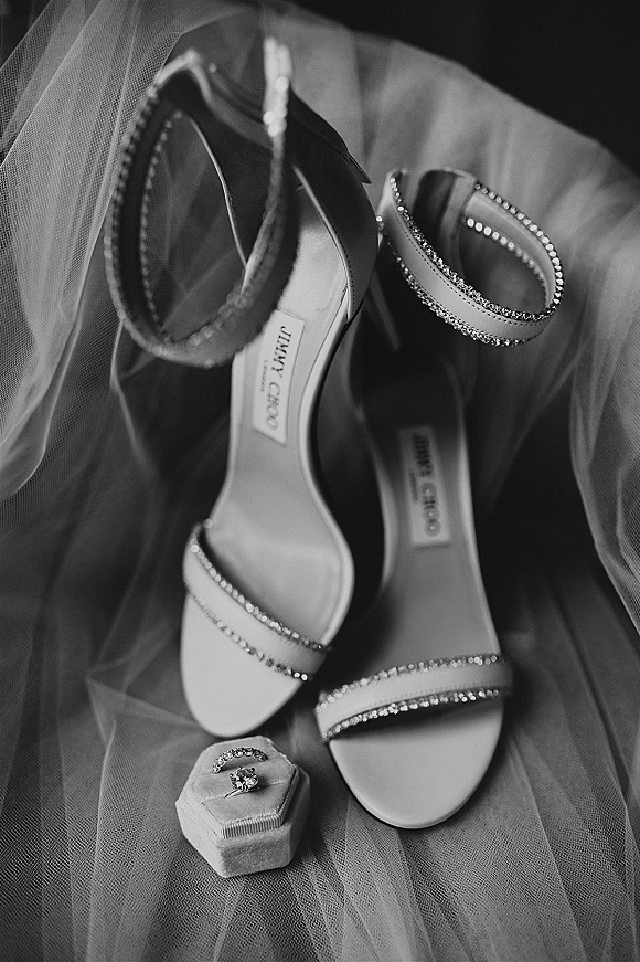 Bridal shoes in a wedding shoe flat lay with rhinestone strap heels beside an engagement ring and ring box on soft tulle fabric backdrop