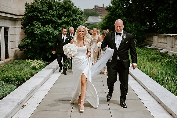 Wedding party walkout as bride and groom stride with white rose bouquet and long veil, bridesmaids and groomsmen on garden walkway by stone building