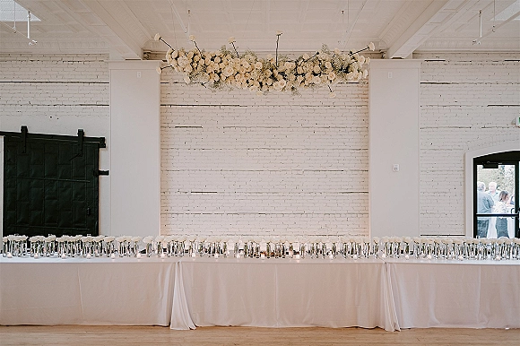 Wedding sweetheart table with a head table floral garland of white roses, bud vases, and candles set against a white brick wall backdrop