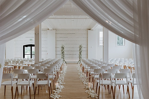 Ceremony setup for an indoor wedding ceremony with white folding chairs, glass candle aisle, rose petals, and draped floral pillars in a bright brick hall