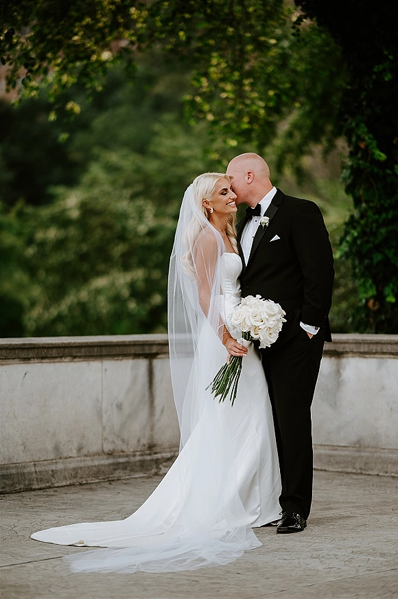 Couple portrait of groom kissing bride’s forehead as she holds a white bouquet, framed by green trees and a stone terrace balustrade