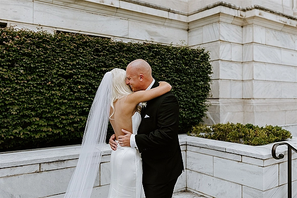 Wedding couple portrait of bride and groom hugging, her veil draped over a strapless satin gown beside an ivy-covered stone wall