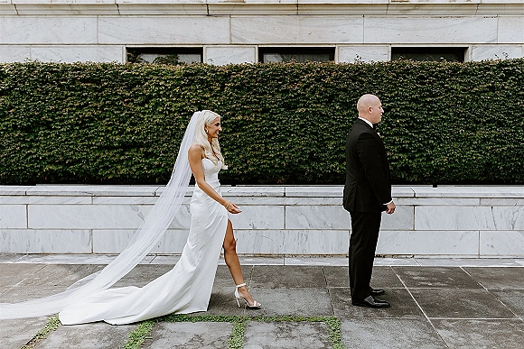 Wedding first look as the bride taps the groom’s shoulder, long cathedral veil trailing along a stone walkway beside an ivy wall